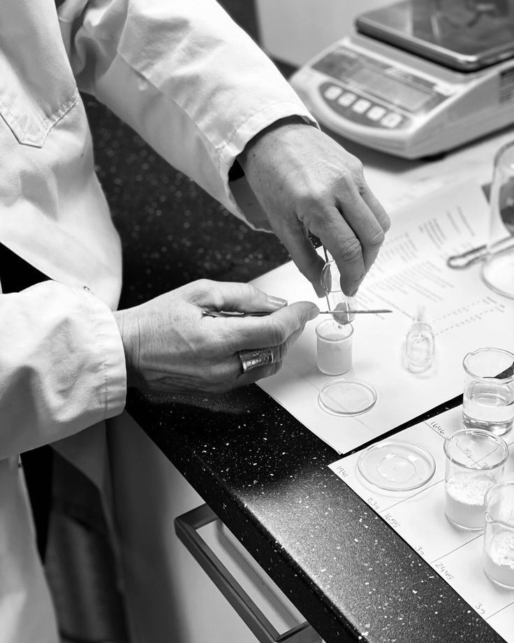 Des mains en blouse de laboratoire pesant une poudre au-dessus d'une balance de précision, entourées de béchers et d'une feuille de pesées.
